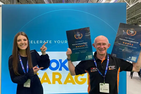 Smiling garage owner holding BookMyGarage excellence certificates in front of a blue branded background.